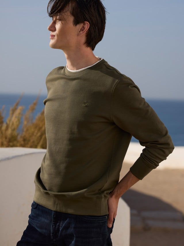 Ein Mann trägt einen olivgrünen Sweatshirt‑Pullover zu dunklen Jeans, was schlicht, modern und entspannt wirkt.