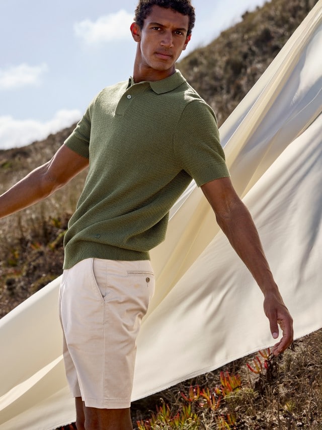 Ein Mann steht auf einem Hügel und trägt ein Poloshirt in Olive, dazu eine helle Shorts, hinter ihm weht ein großes weißes Tuch.