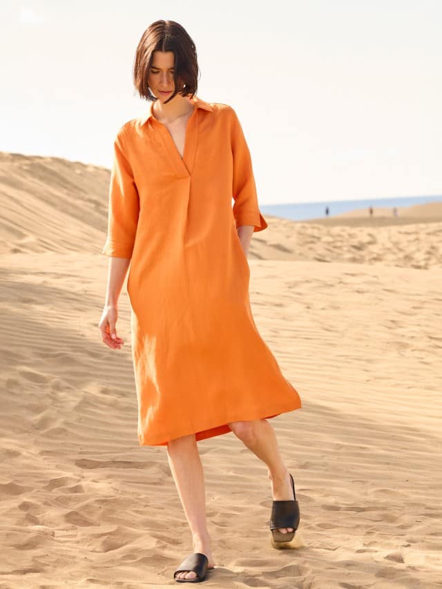 Eine Frau steht in den Dünen und trägt ein Leinenmixkleid in Dunkelorange. Eine Frau steht in den Dünen und trägt ein Leinenmixkleid in Dunkelorange.