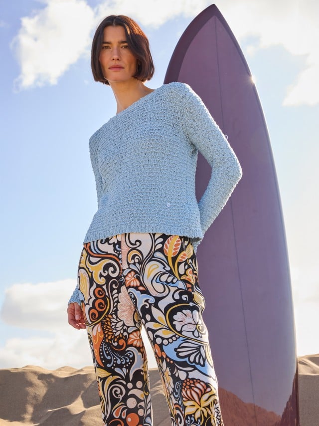 Eine Frau steht vor einem Surfbrett und trägt eine gemusterte Hose, dazu einen Pullover in Hellblau. Eine Frau steht vor einem Surfbrett und trägt eine gemusterte Hose, dazu einen Pullover in Hellblau.