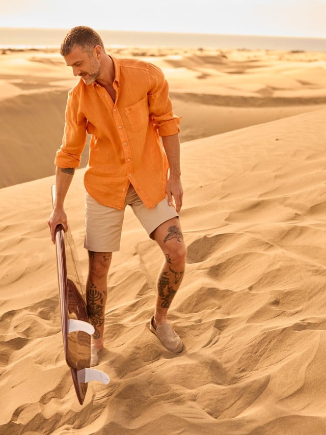 Ein Mann steht mit einem Surfbrett in den Dünen und trägt ein Kurzarmhemd in Orange, dazu helle Shorts.