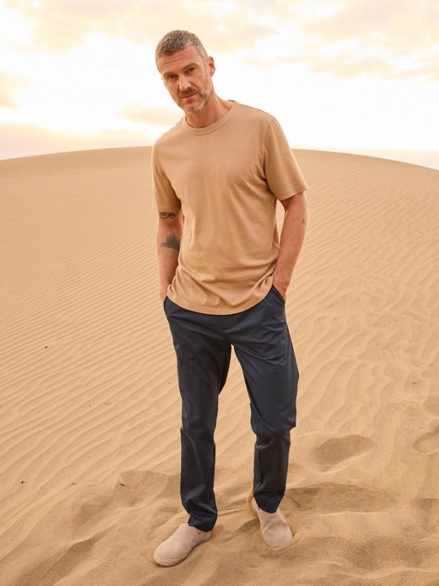 Ein Mann steht in den Dünen und trägt ein helles Shirt, dazu eine Chino in Marine.