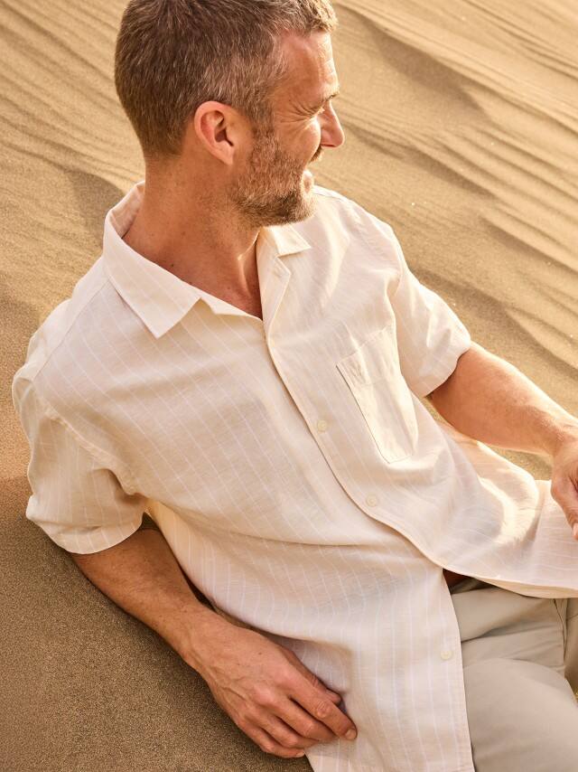 Ein Mann liegt entspannt im Sand einer Düne und trägt ein helles Kurzarmhemd.