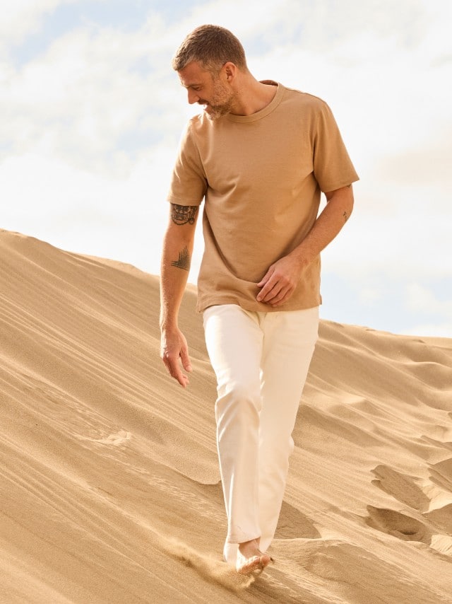 Ein Mann in den Dünen, trägt ein helles Leinen Shirt und dazu eine weiße Hose.