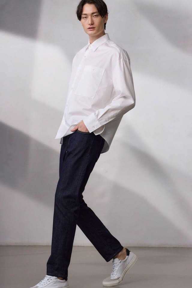 Ein Mann geht vor einem hellen Hintergrund und trägt ein weißes Hemd, dunkle Jeans und weiße Sneaker.