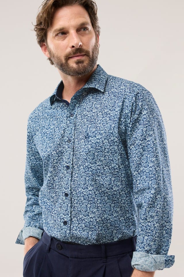 Ein Mann trägt ein blau gemustertes Hemd mit floralem Design und einer dunklen Hose vor neutralem Hintergrund.