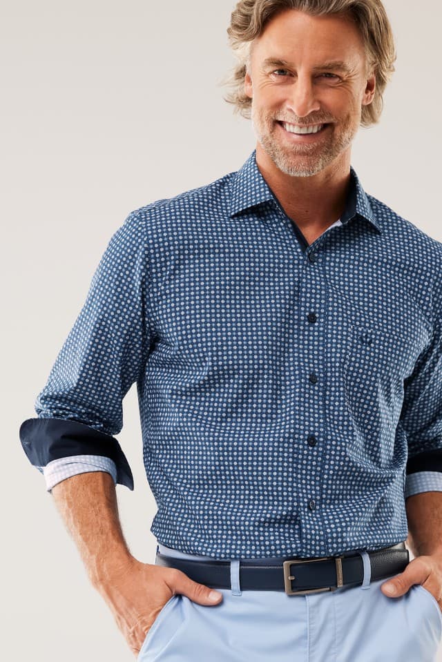 Ein Mann lächelt in die Kamera und trägt ein dunkelblau gemustertes Hemd sowie eine hellblaue Hose mit dunklem Gürtel.