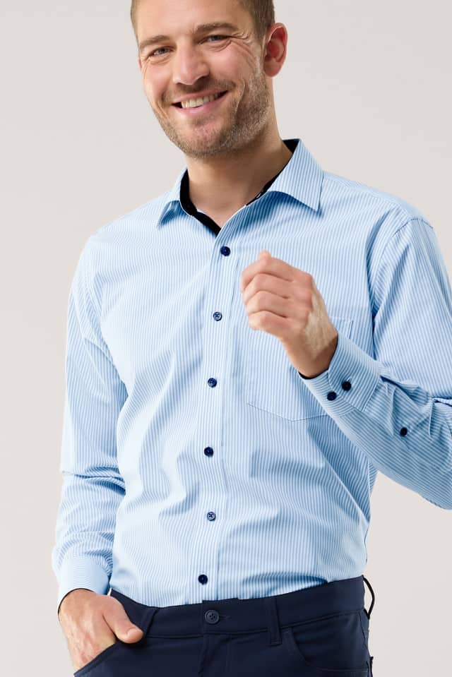 Ein Mann trägt ein hellblau‑weiß gestreiftes Hemd und eine dunkle Hose und lächelt in die Kamera.