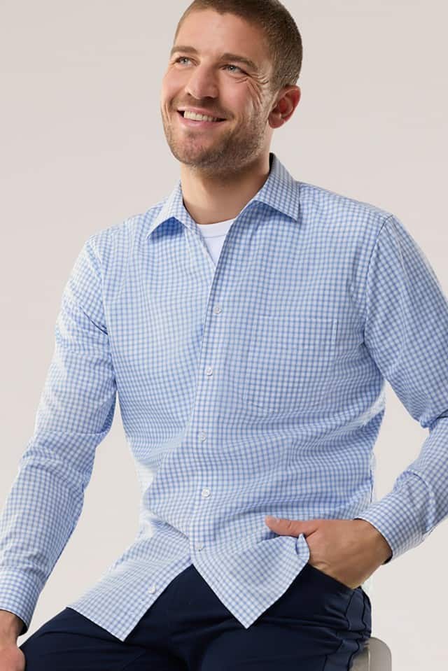 Auf dem Bild ist eine lächelnde Person mit kurz geschnittenem Haar zu sehen, die ein hellblau-weiß kariertes Hemd über einem weißen T-Shirt und eine dunkelblaue Hose trägt.