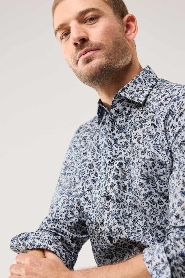 Ein Mann trägt ein helles Hemd mit dunkelblauem floralen Muster vor neutralem Hintergrund.