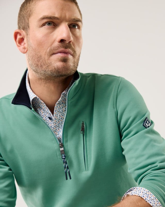 Ein Mann trägt einen grünen Pullover mit kurzem Reißverschluss über einem gemusterten Hemd und blickt zur Seite.