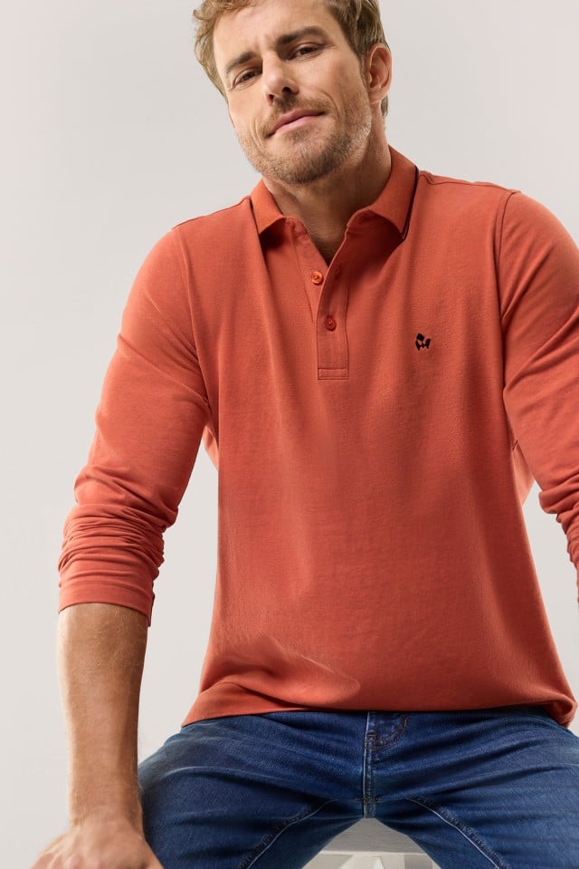 Ein Mann mit kurzem Haar und Bart trägt ein langärmliges, terrakottafarbenes Poloshirt mit dunkelblauer Stickerei auf der Brust und leicht kontrastierender Kragenkante, kombiniert mit einer Jeans.