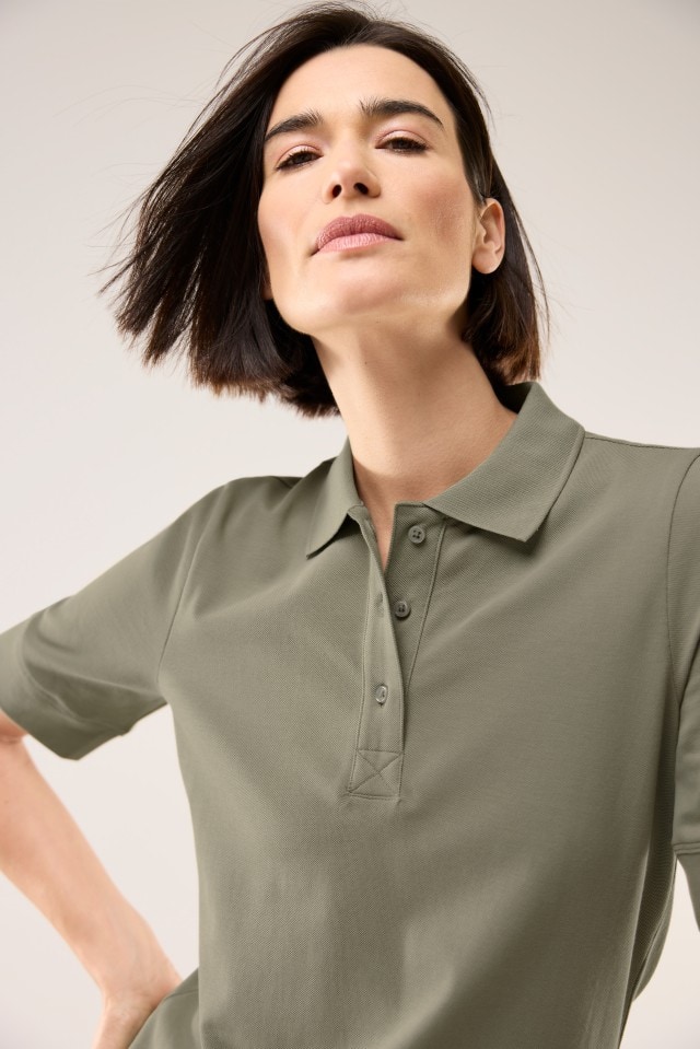 Eine Frau trägt ein olivgrünes Poloshirt mit kurzen Ärmeln und steht vor einem hellen Hintergrund. Die Aufnahme ist nah und zeigt Oberkörper und Gesicht.