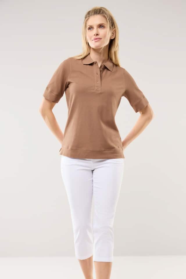 Eine Frau trägt ein kurzärmeliges Poloshirt in Braun und dazu eine weiße, knielange Stretchhose. Sie steht vor einem hellen, neutralen Hintergrund.