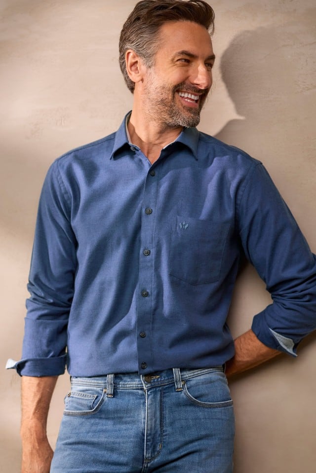 Ein Mann mit grauem Haar und Bart trägt ein dunkelblaues Hemd mit Brusttasche und dunklen Knöpfen sowie eine Jeans und lehnt sich lächelnd an eine helle Wand.