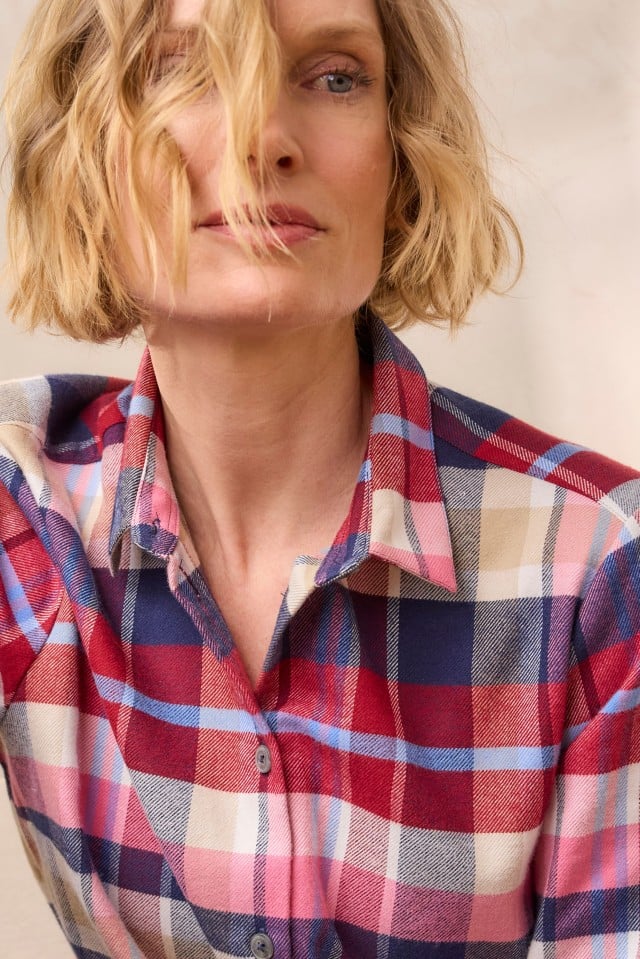 Eine Frau mit blondem, welligem Haar trägt ein kariertes Hemd in Rot-, Blau-, Rosa- und Beigetönen und blickt leicht zur Seite, während ihr eine Strähne ins Gesicht fällt.