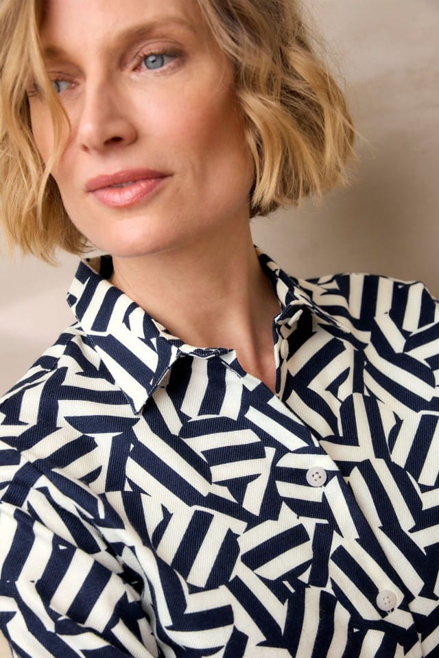 Eine Frau mit blondem, welligem Haar trägt ein Hemd mit auffälligem geometrischem Muster in Dunkelblau und Weiß und blickt leicht zur Seite.