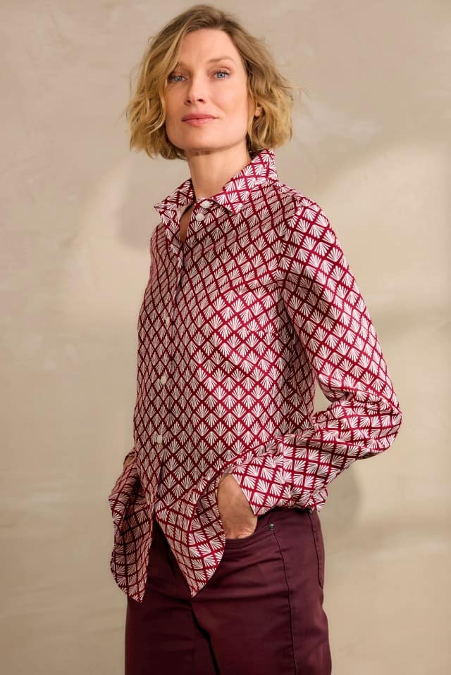 Eine Frau mit blondem, welligem Haar trägt ein Hemd mit rotem und weißem geometrischem Muster sowie eine weinrote Hose und steht mit den Händen in den Taschen vor einer beigen Wand.