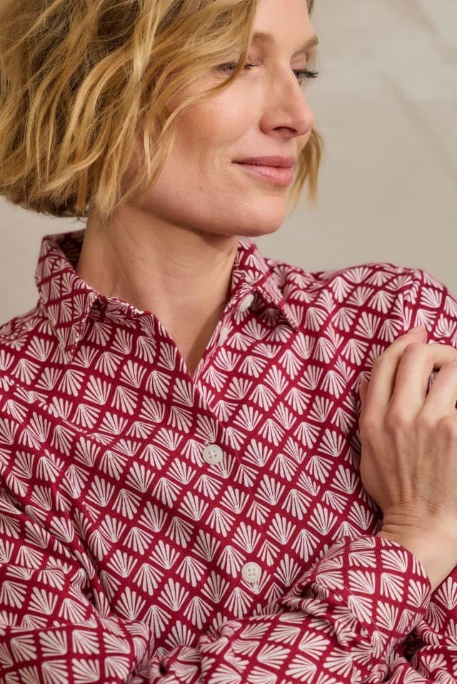 Eine Frau mit blondem, welligem Haar trägt ein rotes Hemd mit weißem, fächerförmigem Muster und blickt seitlich mit einem leichten Lächeln.
