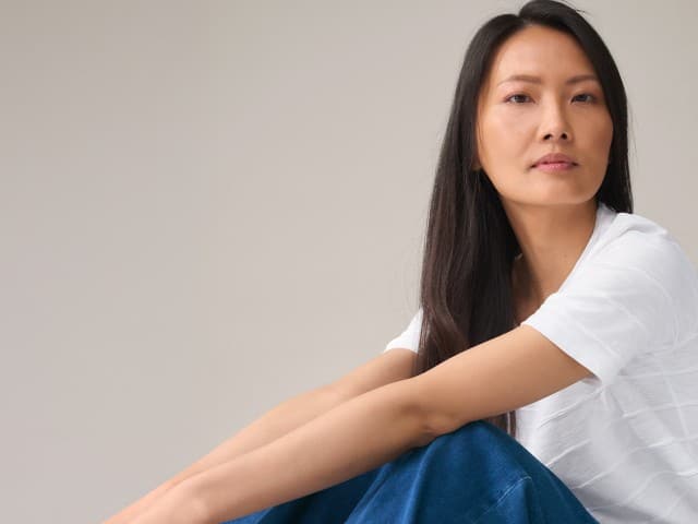 Eine Frau mit langen dunklen Haaren trägt ein weißes T-Shirt und eine blaue Jeans. Sie sitzt mit angewinkelten Beinen und blickt in die Kamera.