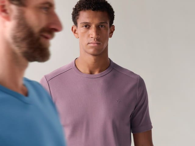 Zwei Männer im Bild: Im Vordergrund ist ein Mann im blauen Shirt unscharf zu sehen, dahinter steht ein Mann in einem lilafarbenen T-Shirt.