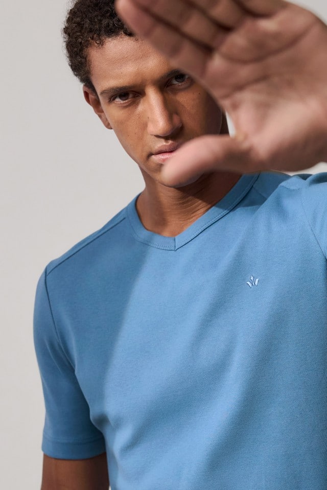 Nahaufnahme eines Mannes mit lockigem Haar in einem hellblauen T-Shirt, der eine Hand vor sein Gesicht hält.

