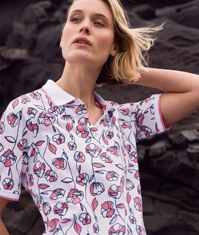 Frau mit blondem Haar in floral gemustertem Poloshirt vor einer dunklen Felslandschaft.