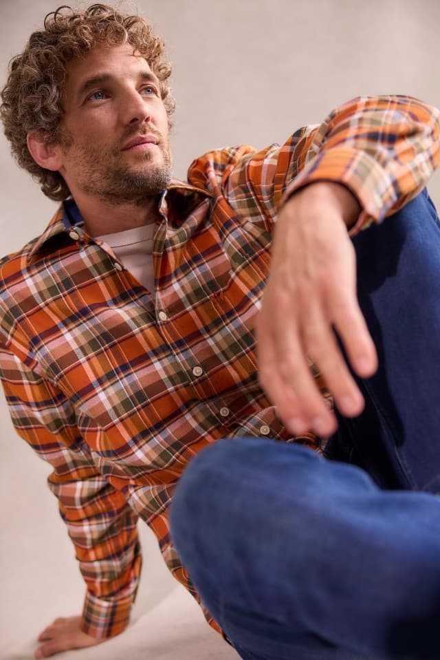 Ein Mann mit lockigem Haar trägt ein orange-braun kariertes Hemd über einem hellen T-Shirt und blaue Jeans, sitzt entspannt und blickt leicht nach oben.