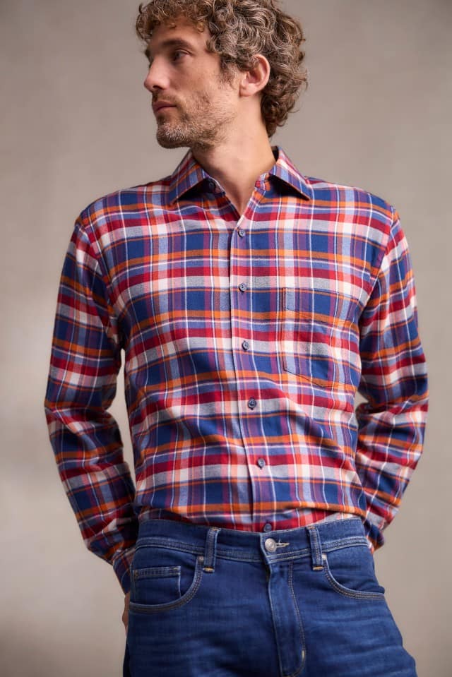 Ein Mann mit lockigem Haar trägt ein bunt kariertes Hemd in Blau-, Rot- und Orangetönen sowie eine blaue Jeans und blickt zur Seite.
