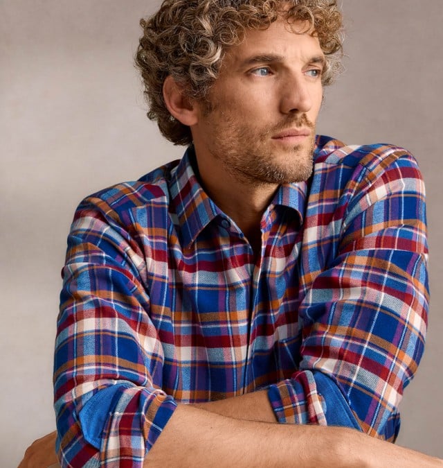 Ein Mann mit lockigem, hellbraunem Haar trägt ein bunt kariertes Hemd in Blau-, Rot- und Brauntönen und blickt nachdenklich zur Seite.