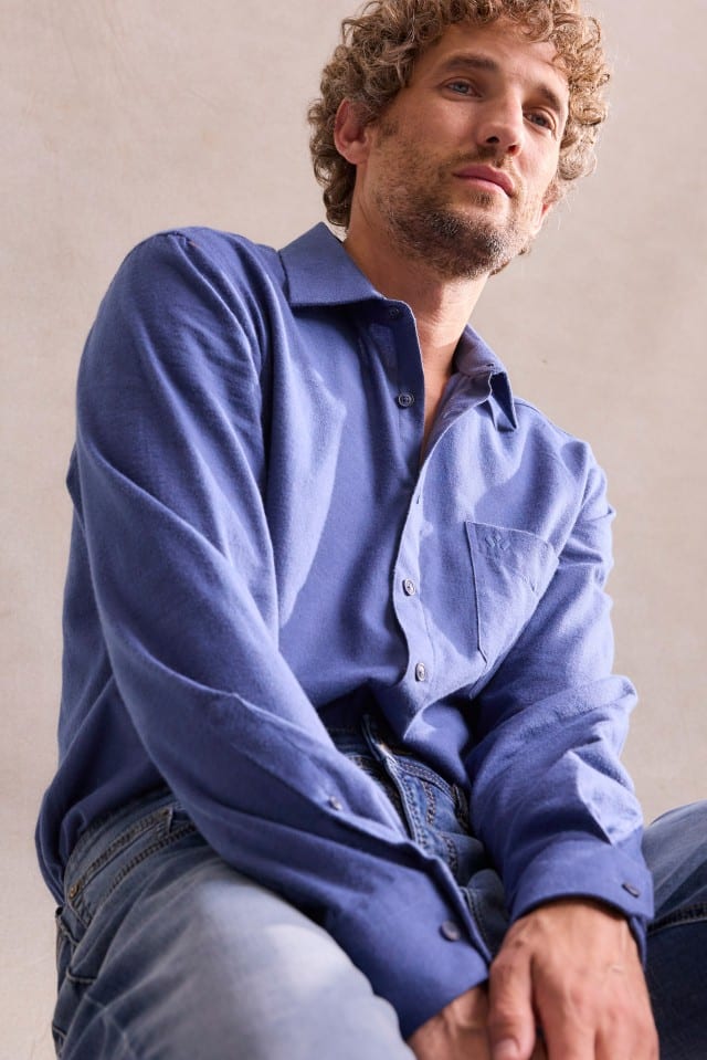 Ein Mann mit lockigem Haar trägt ein blaues Hemd und eine Jeans, sitzt entspannt und blickt leicht nach unten.
