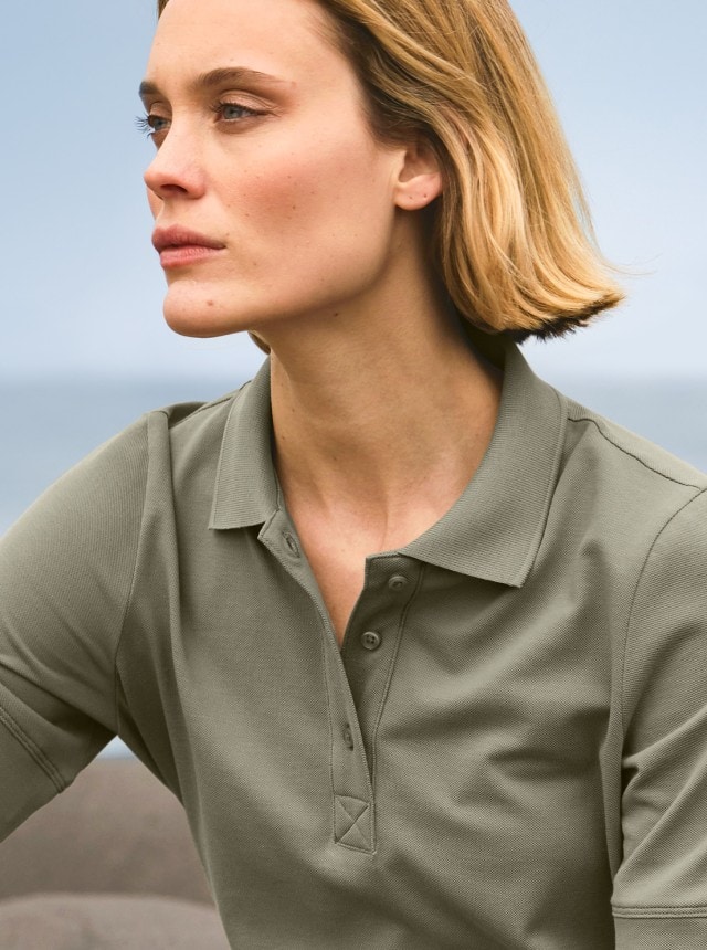 Eine Frau sitzt am Meer und trägt ein olivgrünes Poloshirt.