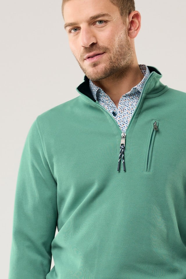 Ein Mann trägt einen grünen Pullover mit kurzem Reißverschluss über einem gemusterten Hemd.