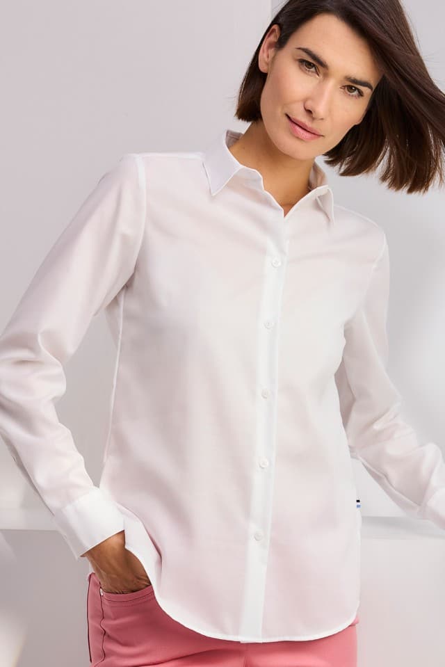 Eine Person mit kinnlangem Haar trägt ein weißes Hemd und rosa Hose und blickt leicht lächelnd in die Kamera.