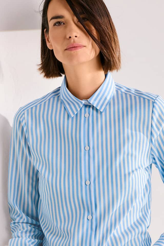 Eine Person mit kinnlangem Haar trägt ein blau‑weiß gestreiftes Hemd und blickt ruhig in die Kamera.