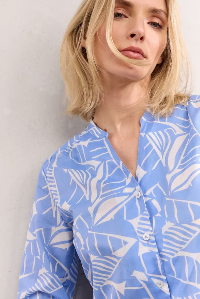 Eine Frau trägt eine hellblau gemusterte Bluse und steht vor einem neutralen Hintergrund.