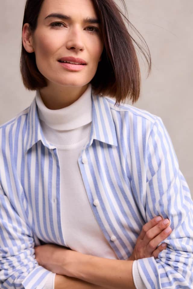 Eine Frau mit schulterlangem, braunem Haar trägt ein weißes Rollkragenshirt unter einer hellblau-weiß gestreiften Bluse und blickt mit leichtem Lächeln in die Kamera.