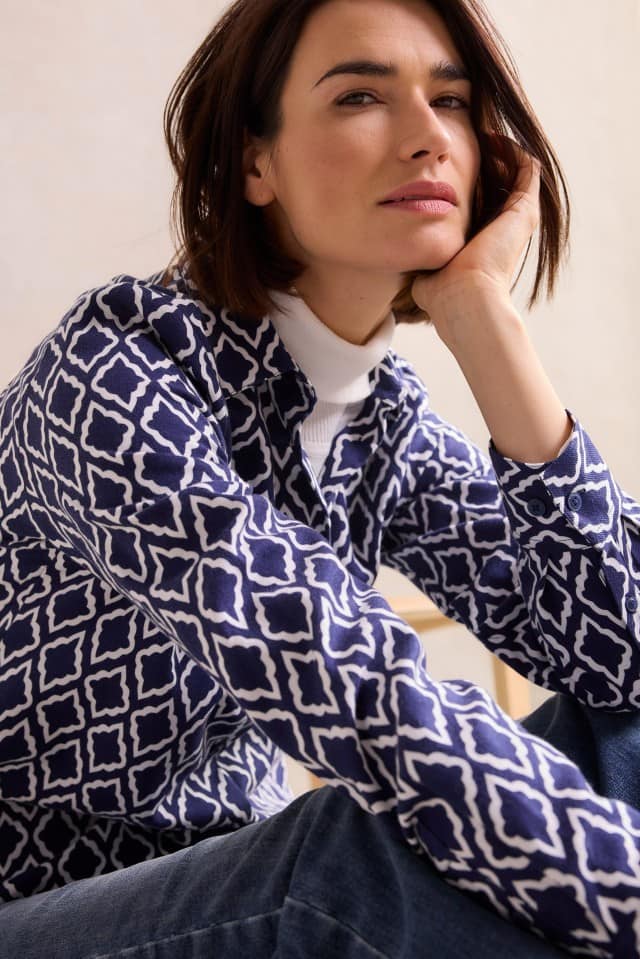 Eine Frau mit schulterlangem, braunem Haar trägt ein weißes Rollkragenshirt unter einer dunkelblauen, geometrisch gemusterten Bluse und stützt den Kopf nachdenklich auf eine Hand.