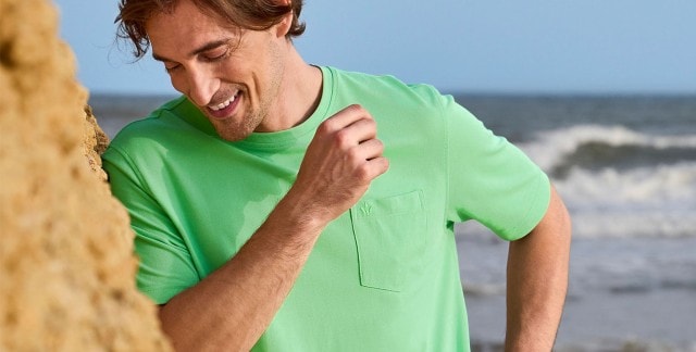 Ein Mann steht an einem Felsen am Meer und trägt ein hellgrünes T-Shirt mit aufgesetzter Tasche auf der Brust.