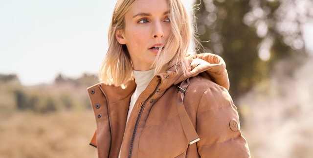 Eine Frau trägt eine hellbraune, wattierte Jacke mit Kapuze, Reißverschluss und breiten Bändern im modernen, schlichten Stil über einem hellen Rollkragenpullover.