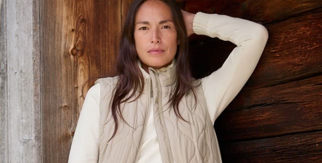 Eine Frau steht vor einer Holzwand und trägt die Wellensteppweste in Toffee/Creme, darunter ein weißes Sweatshirt und dazu eine Jeans. Eine Frau steht vor einer Holzwand und trägt die Wellensteppweste in Toffee/Creme, darunter ein weißes Sweatshirt und dazu eine Jeans.