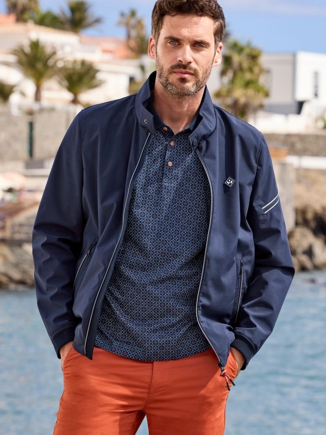 Dunkelhaariger Mann in Club Blouson Marine, Polo-Shirt und Chino in Terra steht an einer Küste, im Hintergrund sind Palmen und Häuser