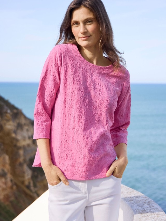 Eine Frau trägt eine pinke Bluse mit Blumenmuster und weiße Hose. Die Kleidung wirkt sommerlich und leger.