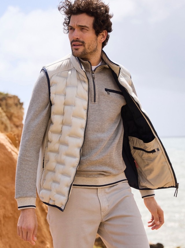 Ein Mann am Strand trägt einen Troyer, darüber eine Steppweste und dazu eine Hose in beige.