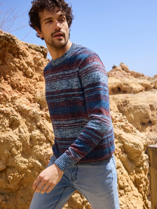 Ein Mann steht an einer Felsküste und trägt einen Strickpullover, dazu eine helle Jeans.