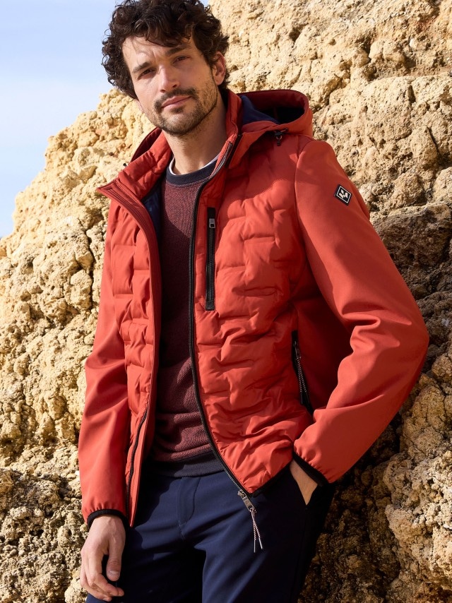 Ein Mann steht vor einer Felswand und trägt einen Blouson in feuerrot, darunter einen roten Pullover und dazu eine dunkele Jeans.