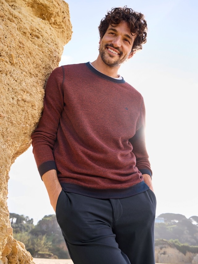 Ein Mann steht vor einer Felswand und trägt einen Struktur Pullover in dunkelrot, dazu eine Hose in marine.