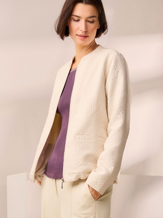 Eine Frau trägt eine cremefarbene, gesteppte Jacke mit dezentem Blumenmuster über einem gerippten, violetten Oberteil, kombiniert mit einer hellen Hose im eleganten, modernen Stil.