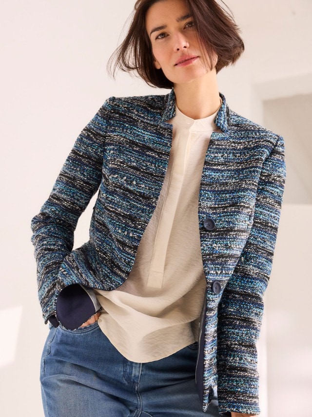 Eine Frau trägt einen strukturierten Blazer in Blau-, Grau- und Schwarztönen über einer cremefarbenen Bluse, kombiniert mit einer Jeans im stilvoll-lässigen Look.
