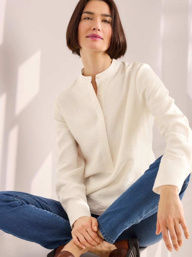 Eine Frau trägt eine cremeweiße Bluse mit kleinem Stehkragen und verdeckter Knopfleiste, kombiniert mit einer blauen Jeans und braunen Loafern im klassischen, lässigen Stil.